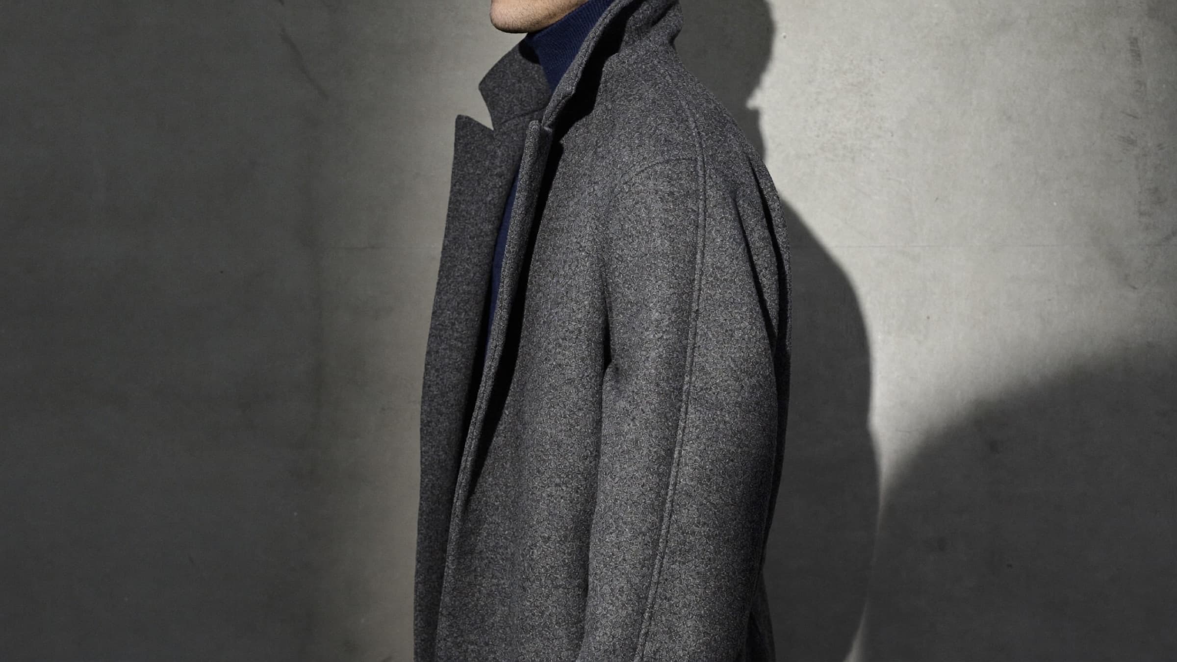 Homme de dos en manteau raglan de laine gris anthracite, dans une pièce en béton brut